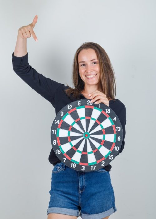 Young woman pointing finger at dartboard in black shirt, jeans shorts and looking cheerful , front view.